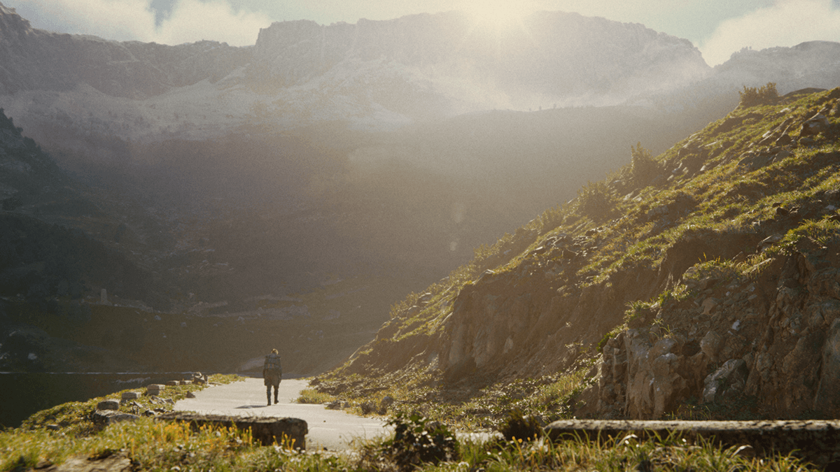 A figure in a military gear walks alone along a mountain road.