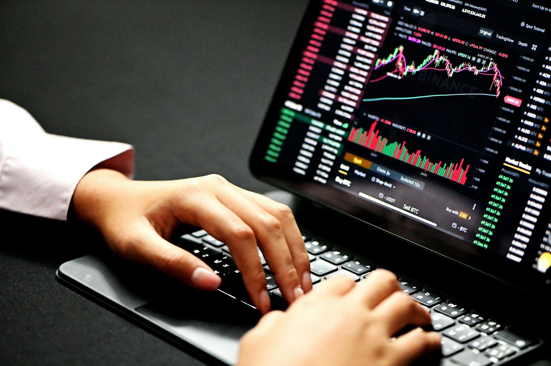 A pair of hands resting on a keyboard with an iPad showing graphs and price quotes. (Kanchanara/Unsplash)