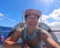 A woman with a sunglasses tan, wearing a tank top and bandana and watch, taking a selfie on a small craft, surrounded by water.