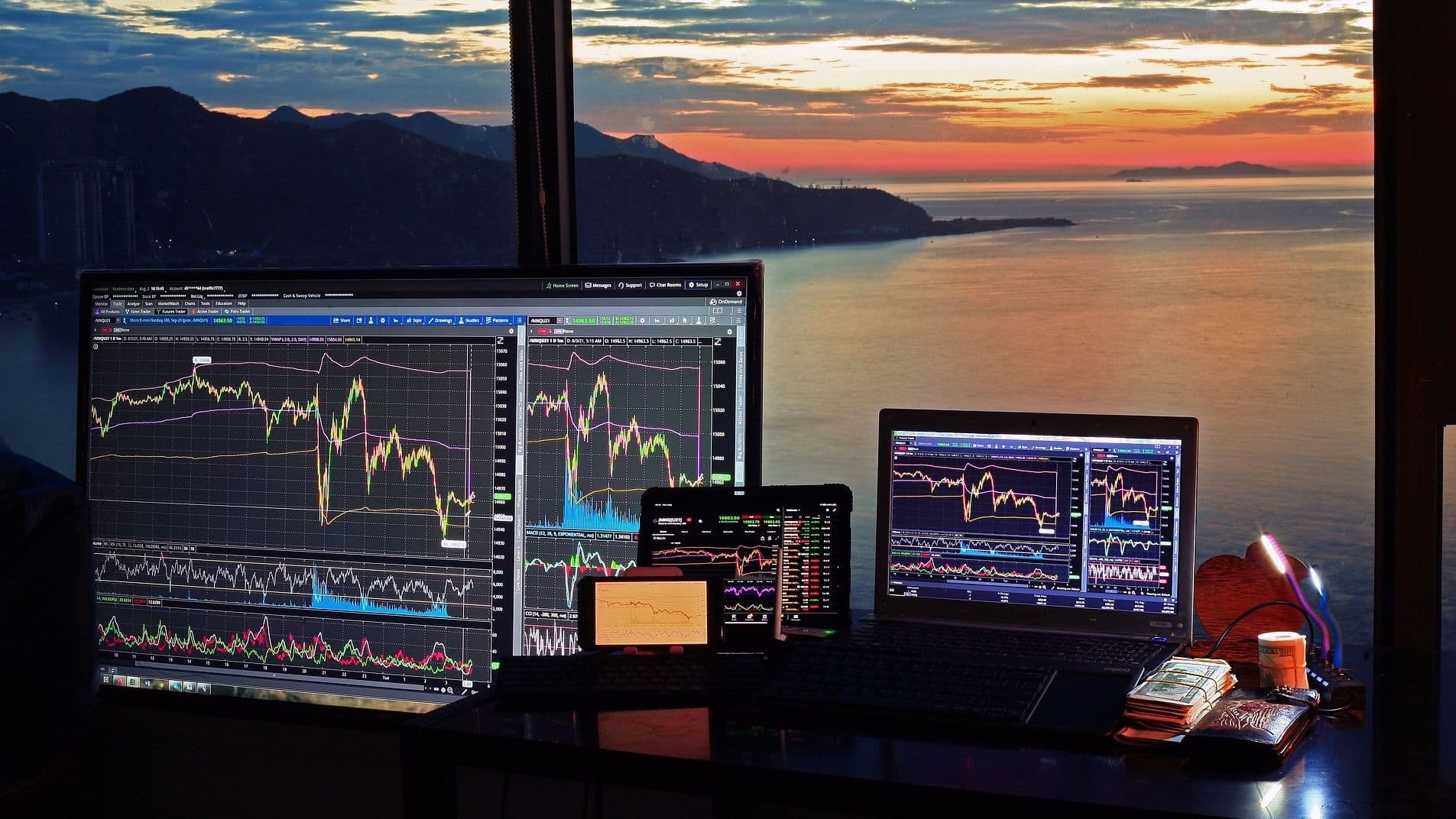 Computer monitors and a laptop screen show trading charts on a desk overlooking an expanse of water at sunset. (sergeitokmakov/Pixabay)