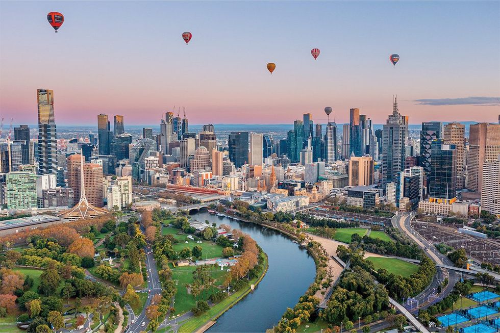 Hot-air balloons over a riverside city skyline at sunrise with parks in the foreground