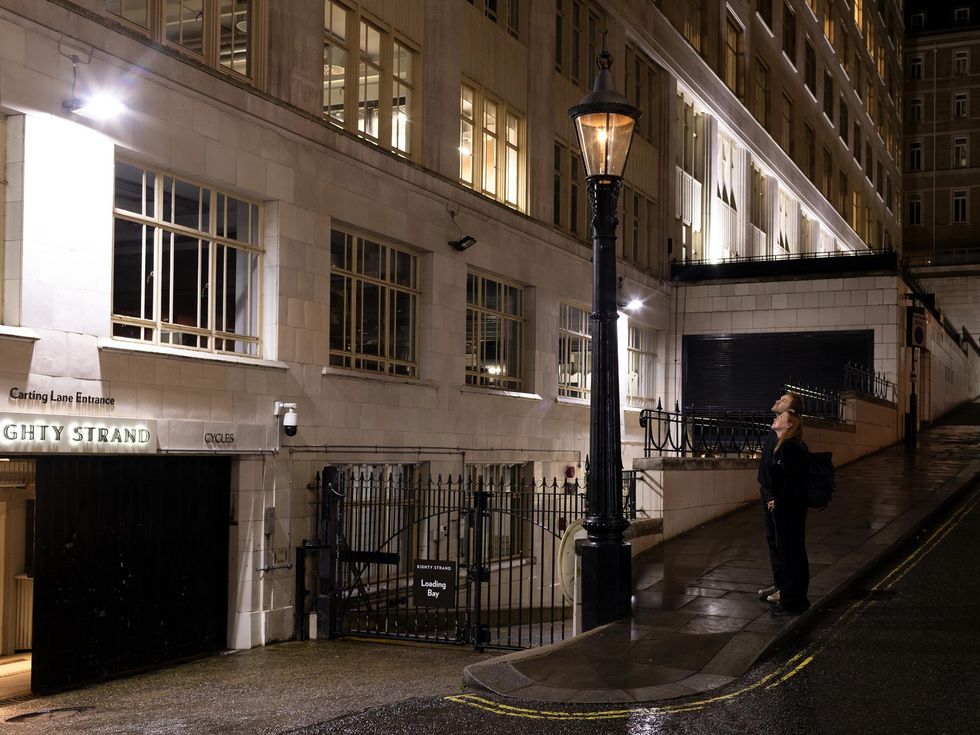 Two tourists observe the faint flame of the historic Webb Patent Sewer Gas Lamp on Carting Lane, with bright white LED lighting from modern fixtures nearby.