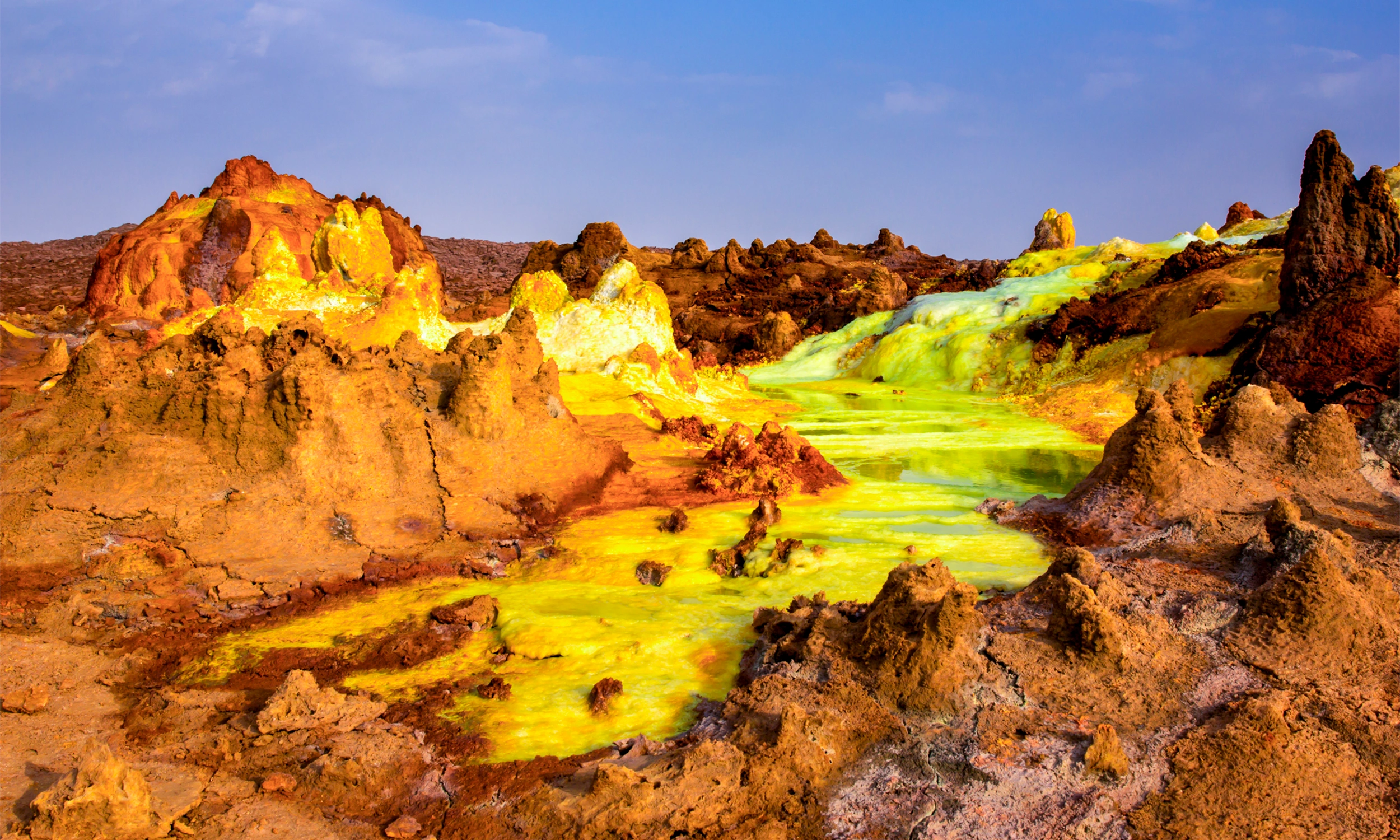 Vividly colored mineral terraces in Ethiopia’s Danakil Depression in yellow, green and orange hues, with pools of dark water nestled among the crusted formations, under a pale sky