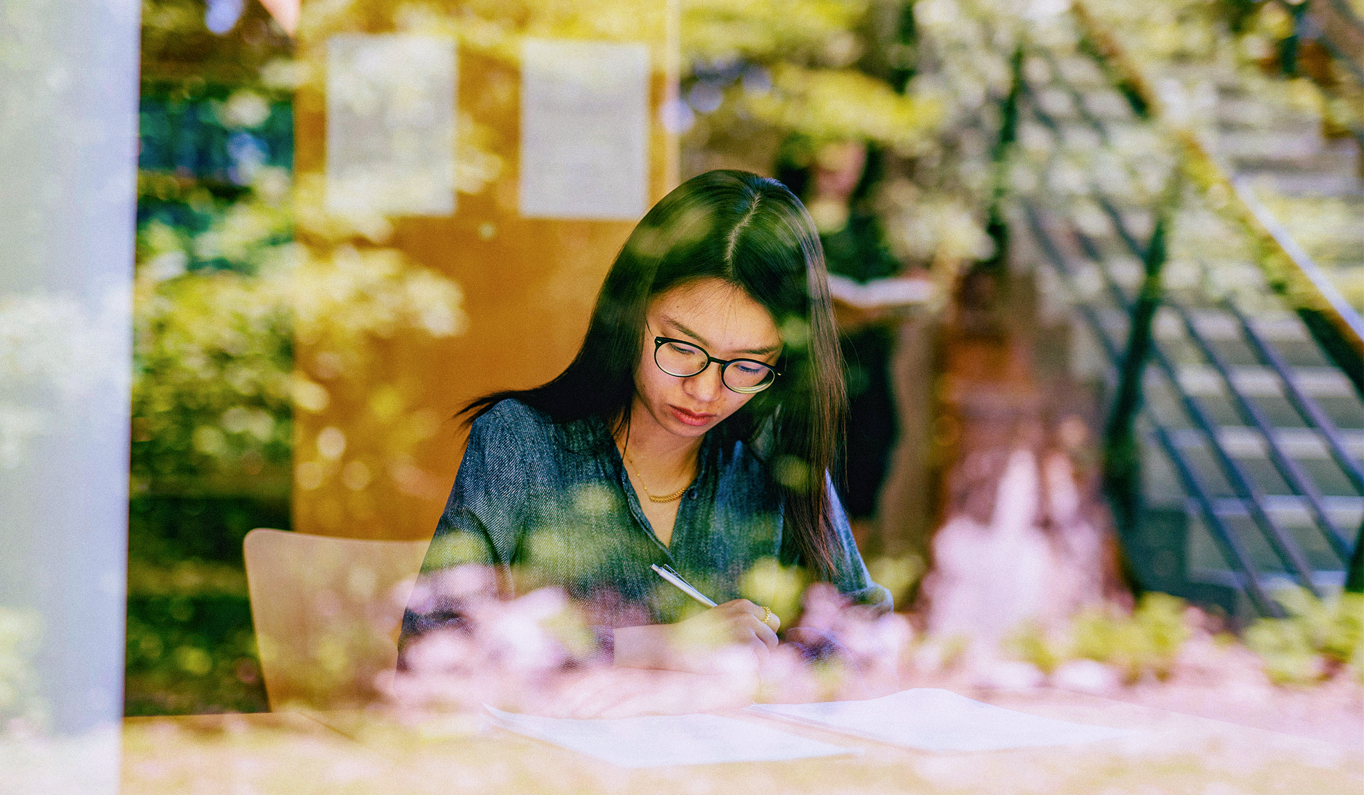 A woman seated in a library, looking down at her work, as seen through a window with reflections of greenery and flowers.