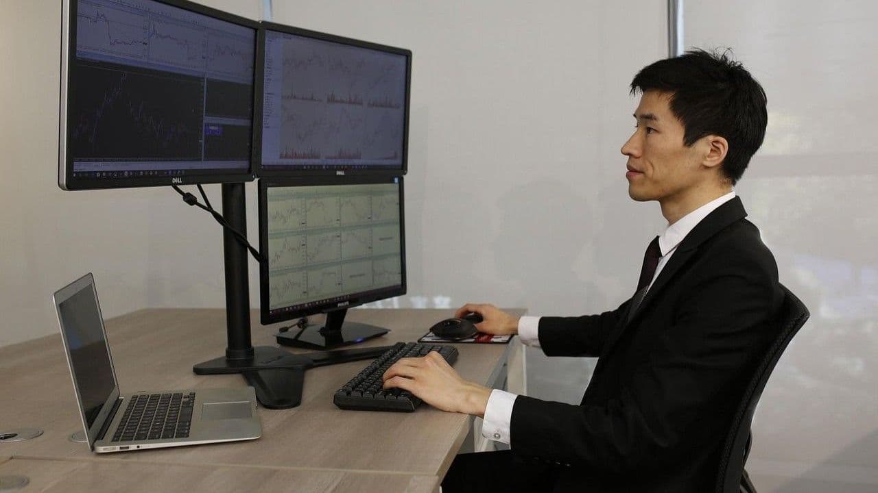 A besuited trader monitors three desktop screens and a laptop. (harryloya/Pixabay)