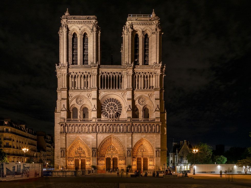 The west facade of the Notre-Dame cathedral glows at night following restoration, with warm white architectural LED lighting illuminating the three monumental portals, rose window, and twin towers.