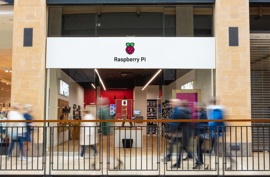 The image depicts the exterior of a Raspberry Pi store. Here are the key details: Store Details: The store prominently displays the Raspberry Pi name and logo above its entrance. Through a large glass window, we can glimpse the well-lit interior with various items on display. Blurred figures of people are seen walking in front of the store, suggesting motion. A metal railing separates the walking area from a lower level in the mall. The architecture features beige-colored walls and pillars. Raspberry Pi: The store specializes in products related to Raspberry Pi, a popular single-board computer used for various projects and educational purposes.