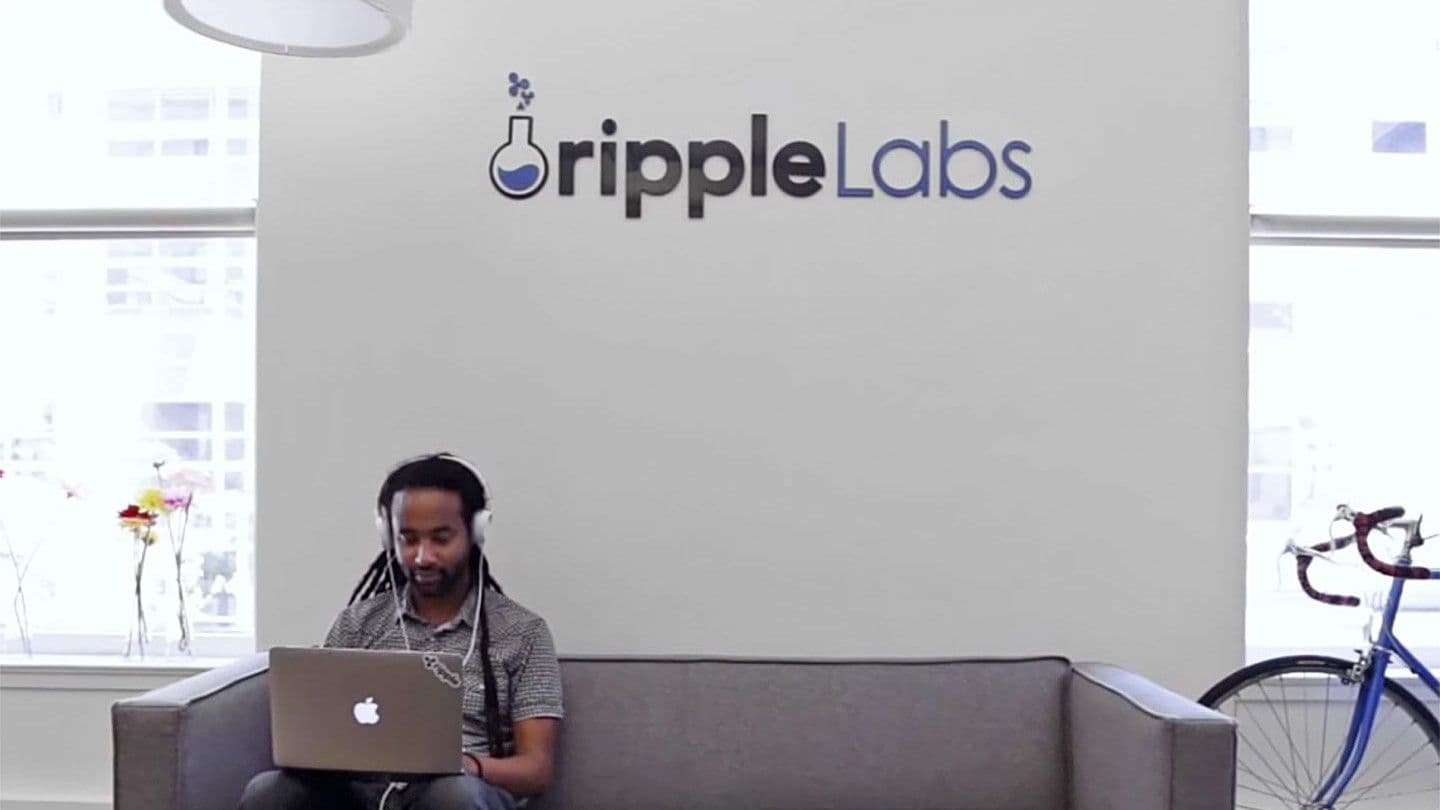 A man seated on a sofa works on a laptop under a Ripple Labs sign. (Ripple Labs)